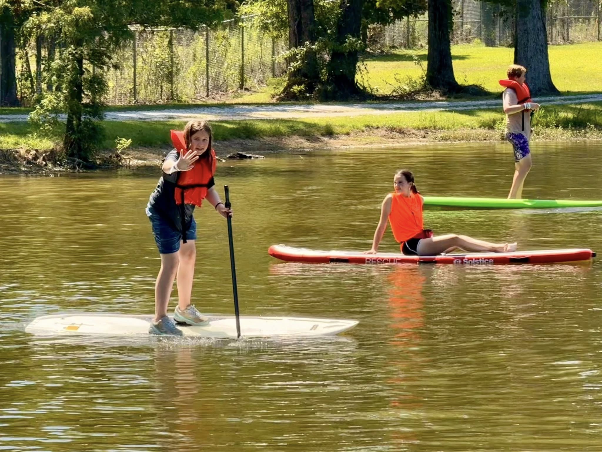 Three people paddleboarding on a calm lake, wearing life vests; one waves, another sits, and one stands.